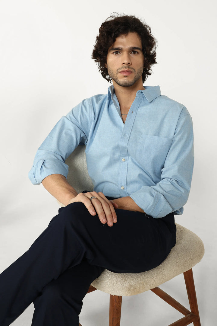 Man wearing a light blue shirt and dark pants sitting on a chair against a white background