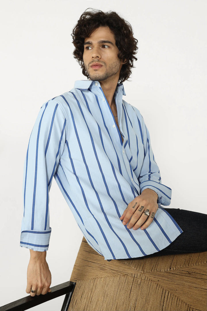 Man wearing a light blue striped shirt sitting on a wooden chair against a white background