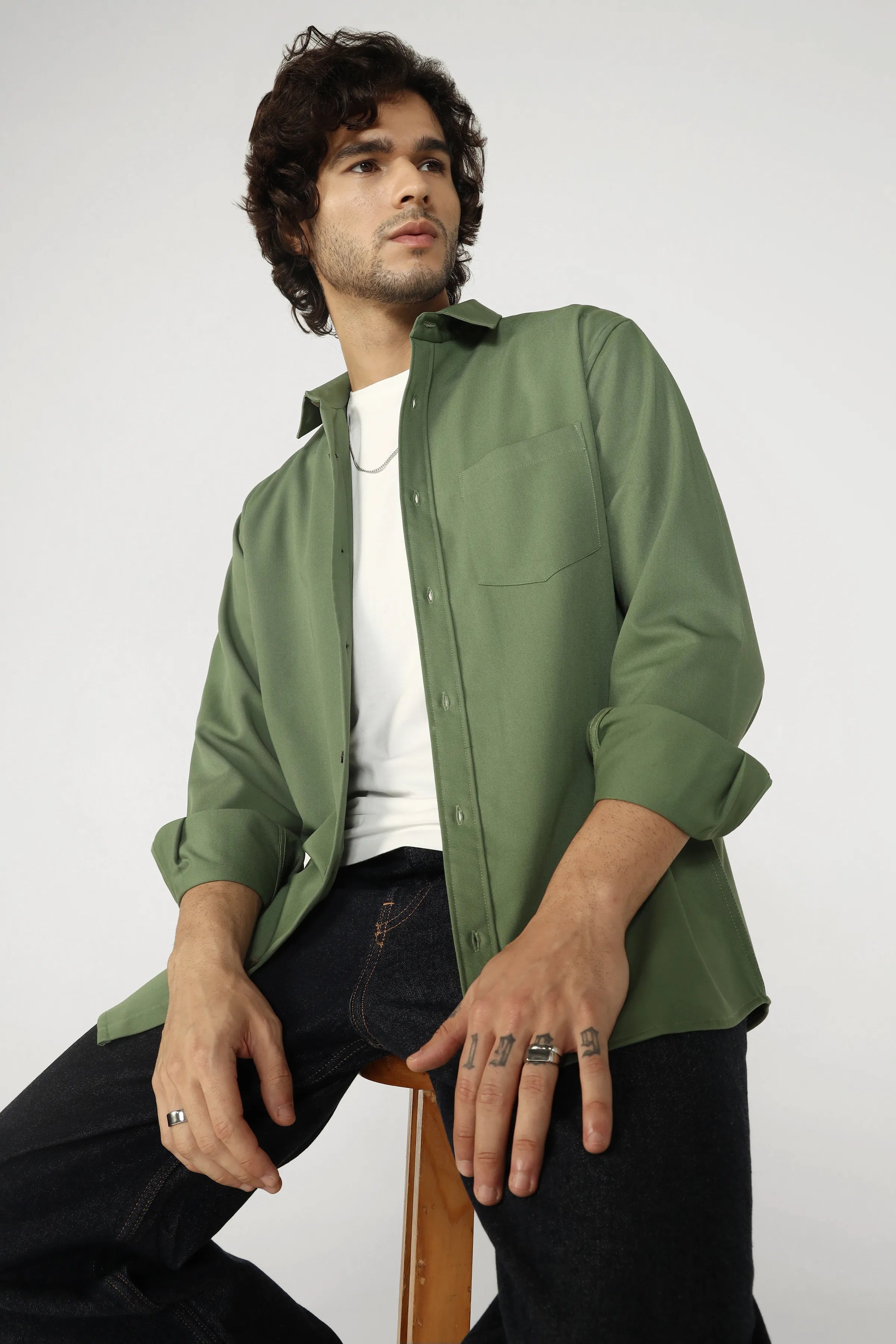 Man wearing a green shirt over a white t-shirt, sitting on a wooden stool against a plain background