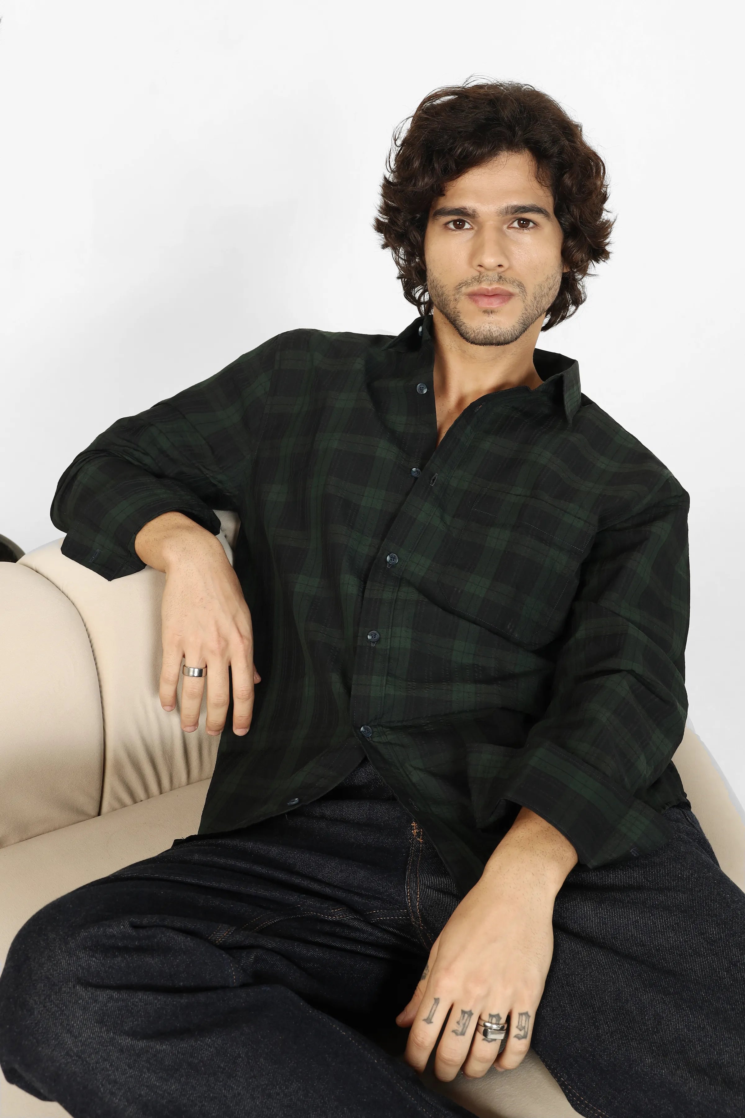 Man sitting on a couch wearing a green seersucker shirt and dark pants against a white background