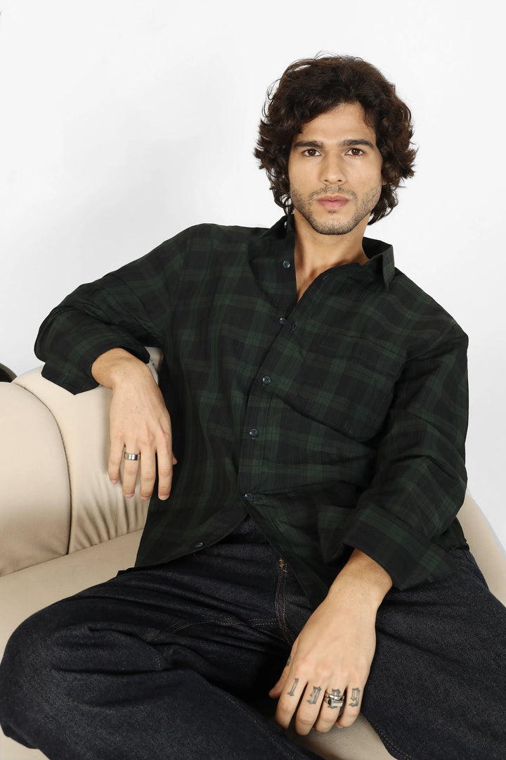 Man sitting on a couch wearing a green seersucker shirt and dark pants against a white background
