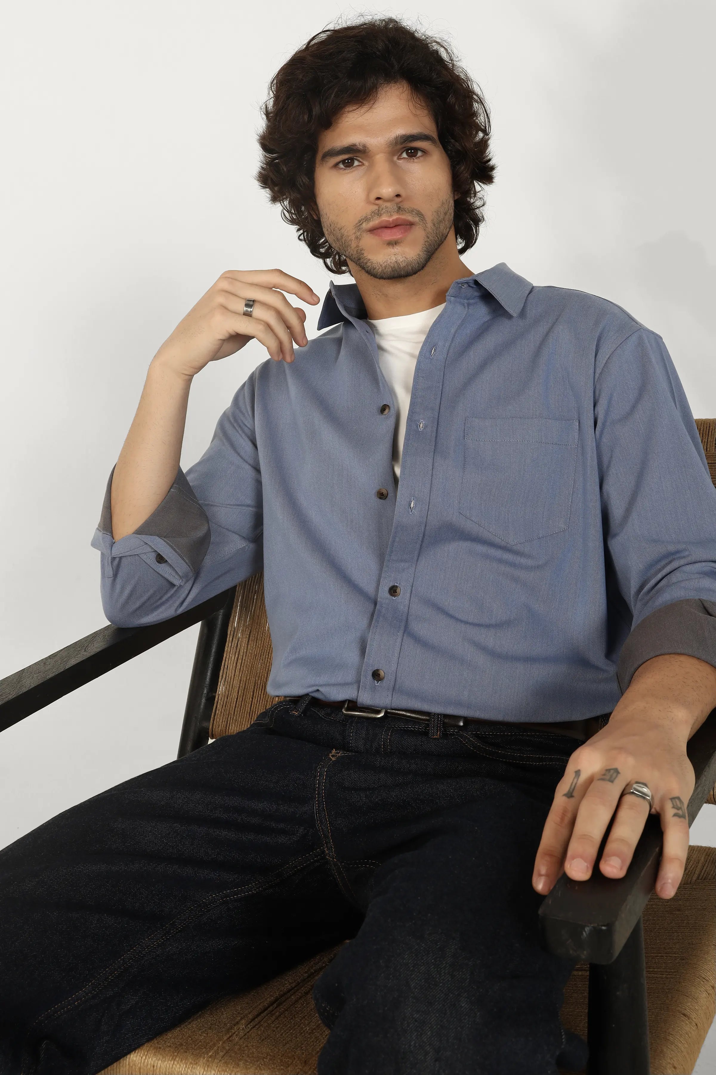 Man wearing a Oxford Cotton Twill Shirt and dark jeans sitting on a chair against a white background