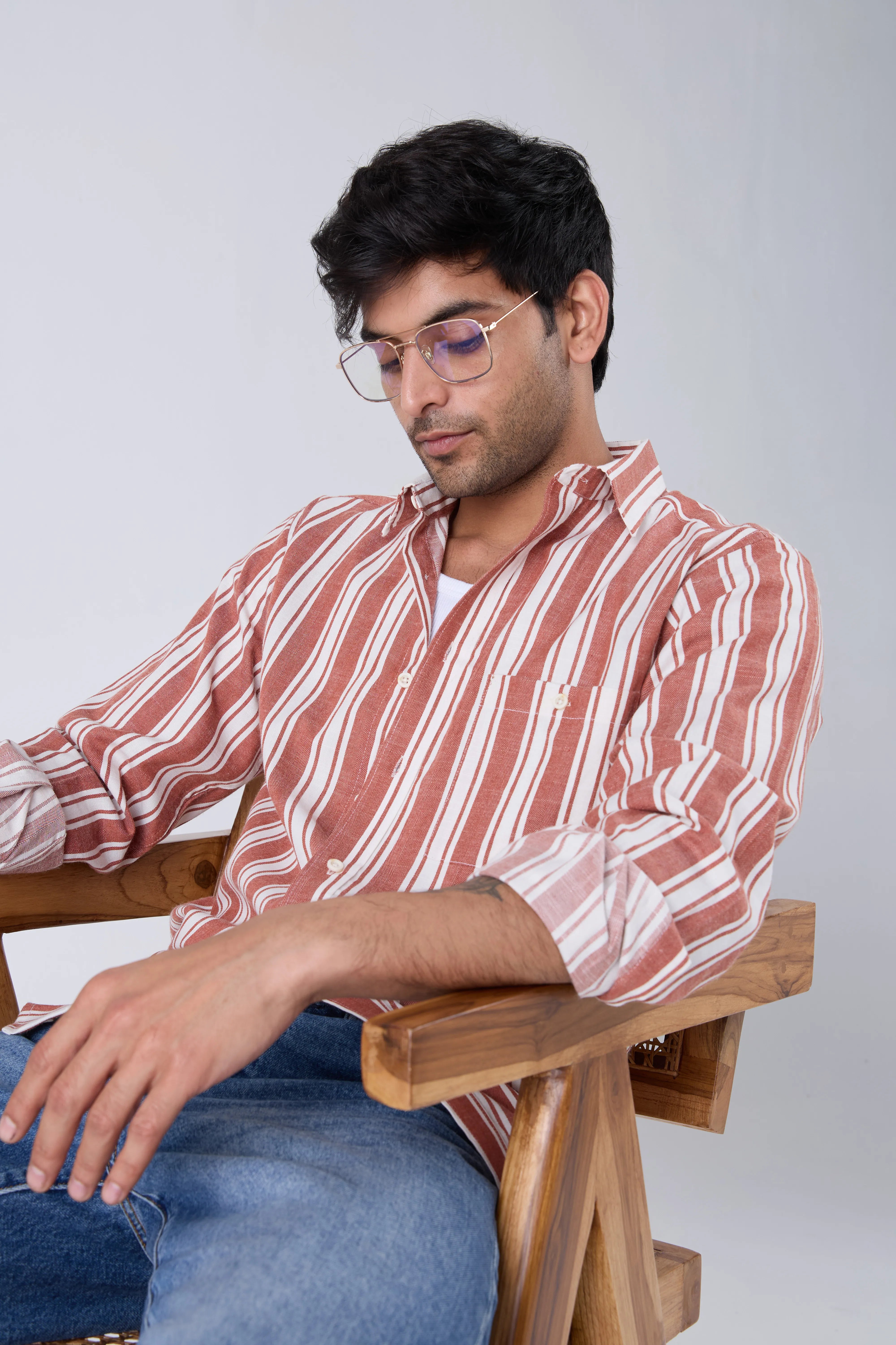 Man wearing a tan cotton striped shirt and glasses sitting on a wooden chair against a plain background